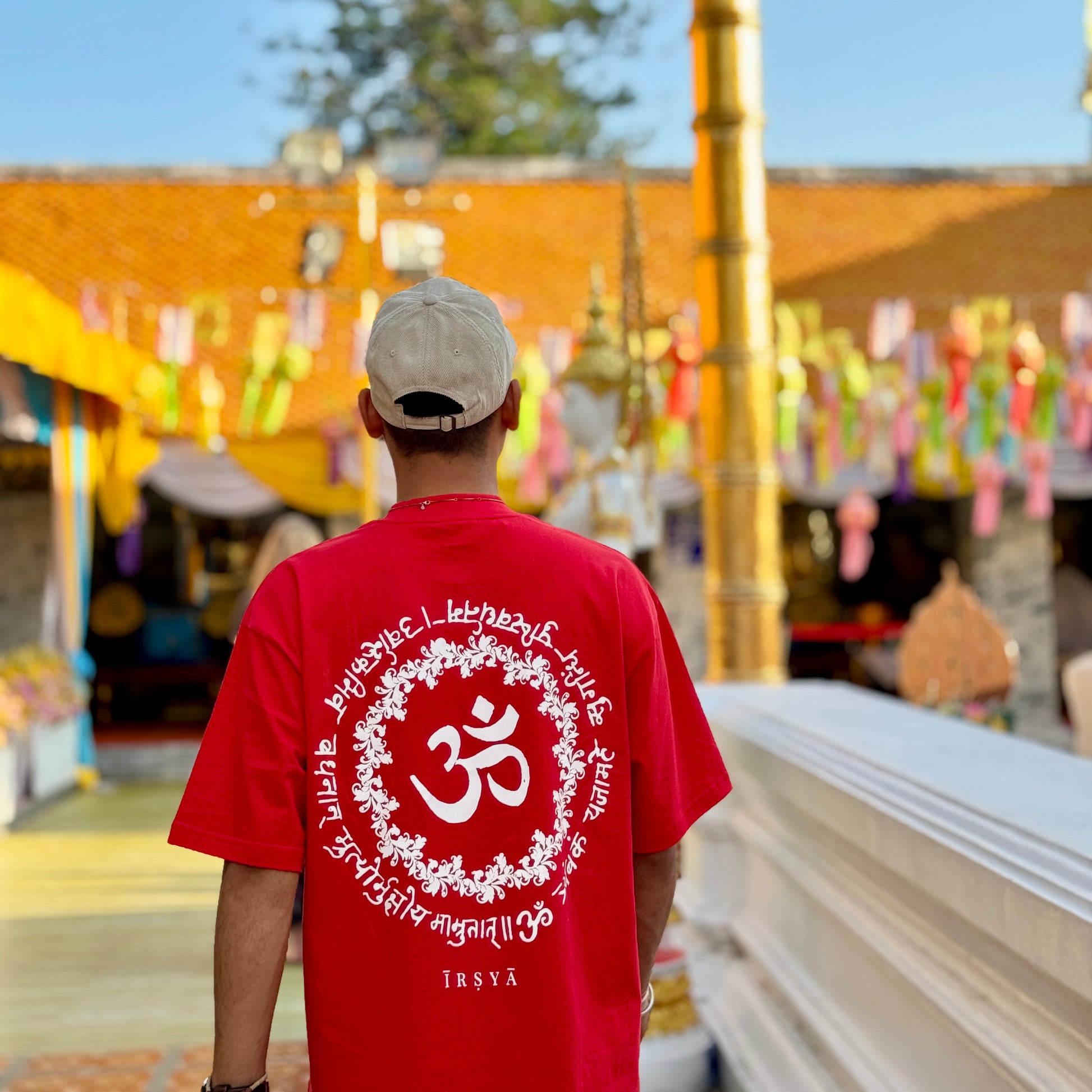 A person wearing a red oversized t-shirt with a white spiritual mantra screen print, standing in front of a colorful backdrop.